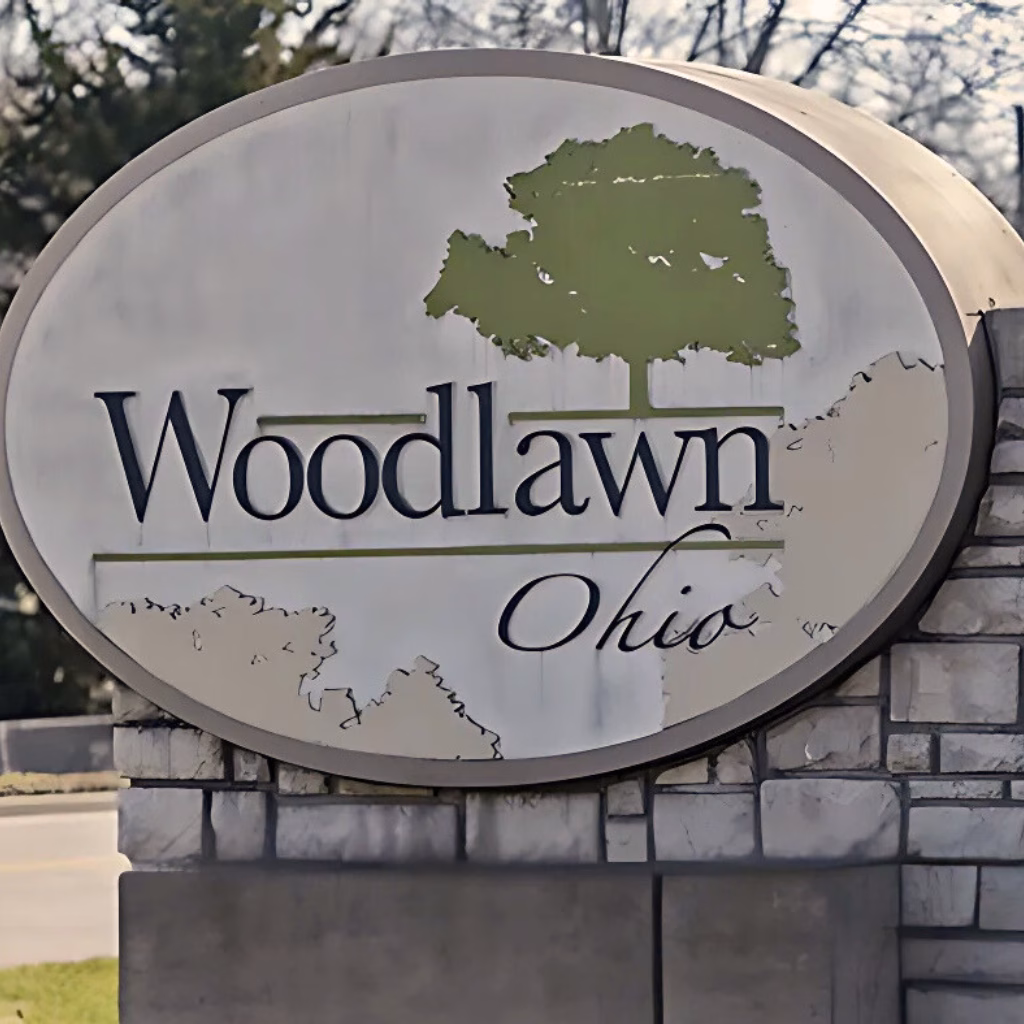 A close-up of the Woodlawn, Ohio, welcome sign framed by stonework and a tree, highlighting a peaceful suburban setting where many residents search for a dispensary near Woodlawn for trusted cannabis access.
